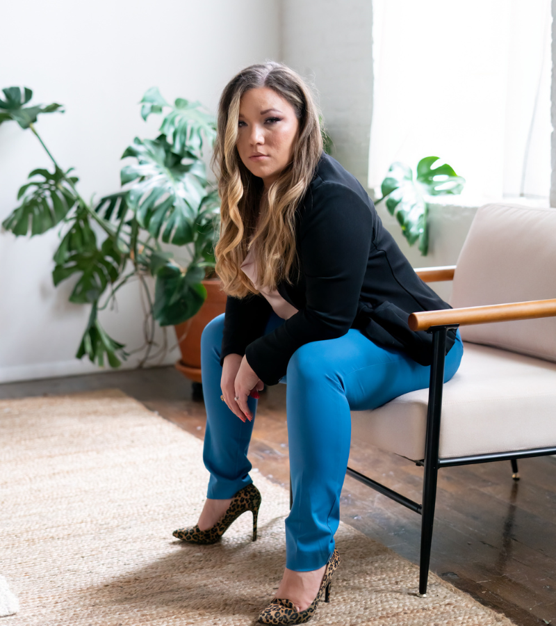 Ashley Marie sitting in chair with blue pants, black top, blonde hair and leopard shoes with a serious face